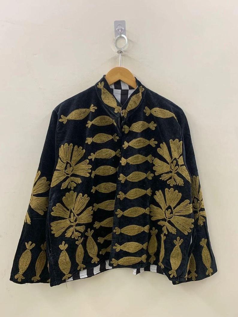 May include: Black jacket with gold floral and fish-shaped embroidery. The jacket has a high collar and a zipper closure. The lining is black and white checkered. The jacket is hanging on a wooden hanger.