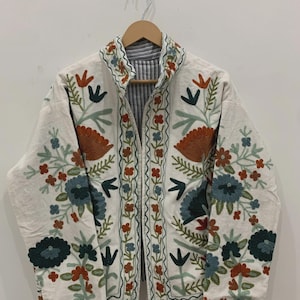 Puede incluir: Chaqueta blanquecina con bordados florales coloridos. La chaqueta presenta un cuello mandar&iacute;n y un forro a rayas. El bordado incluye flores y hojas naranjas, azules y verdes. La chaqueta est&aacute; colgada de una percha de madera.