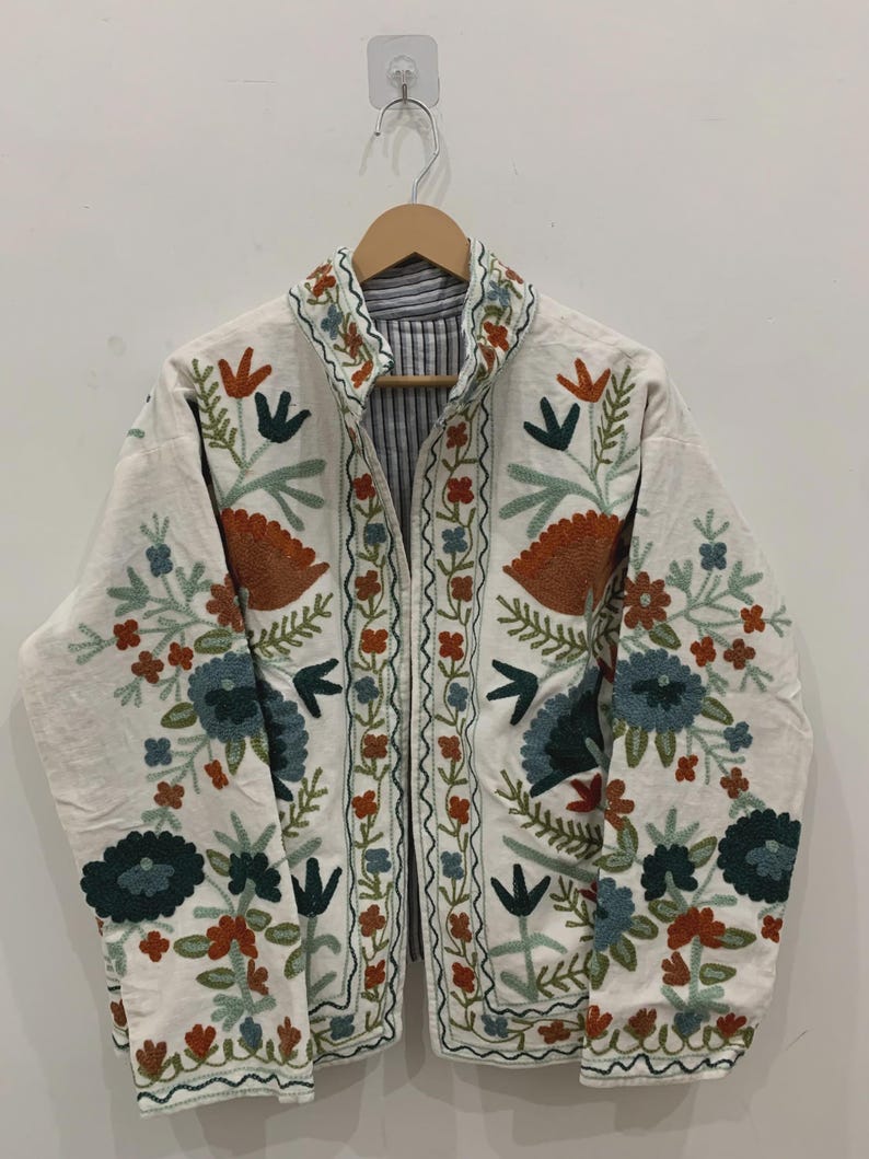 Puede incluir: Chaqueta blanquecina con bordados florales coloridos. La chaqueta presenta un cuello mandar&iacute;n y un forro a rayas. El bordado incluye flores y hojas naranjas, azules y verdes. La chaqueta est&aacute; colgada de una percha de madera.