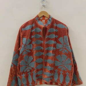 May include: Rust-colored jacket with blue floral embroidery. The jacket features a mandarin collar and long sleeves. The embroidery includes large flowers and leaf-like shapes. The jacket is hanging on a wooden hanger against a white background.
