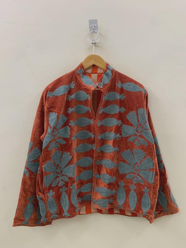 May include: Rust-colored jacket with blue floral embroidery. The jacket features a mandarin collar and long sleeves. The embroidery includes large flowers and leaf-like shapes. The jacket is hanging on a wooden hanger against a white background.
