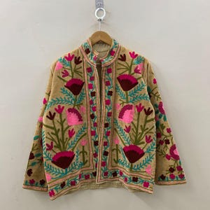 Puede incluir: Chaqueta beige bordada con un diseño floral en tonos rosa, burdeos y turquesa. La chaqueta tiene cuello mandarín y mangas largas. El bordado es detallado y cubre la parte delantera, trasera y mangas de la chaqueta.