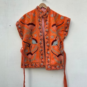 May include: An orange embroidered vest with floral patterns in teal, burgundy, and brown. The vest has a high collar and tie closures. The interior lining features a red and white striped pattern. The vest is hanging on a wooden hanger.