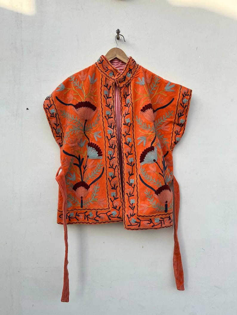 May include: An orange embroidered vest with floral patterns in teal, burgundy, and brown. The vest has a high collar and tie closures. The interior lining features a red and white striped pattern. The vest is hanging on a wooden hanger.