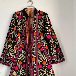 May include: Black embroidered jacket with floral patterns in gold, orange, and pink. The jacket has a striped interior and is hanging on a silver hanger. The jacket's design features a collar and three-quarter sleeves.