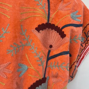 May include: Close-up of an orange embroidered garment. The garment features a floral design with pink, blue, and yellow thread. The embroidery is detailed and covers a large portion of the fabric. The garment's edge has a red and white striped trim.