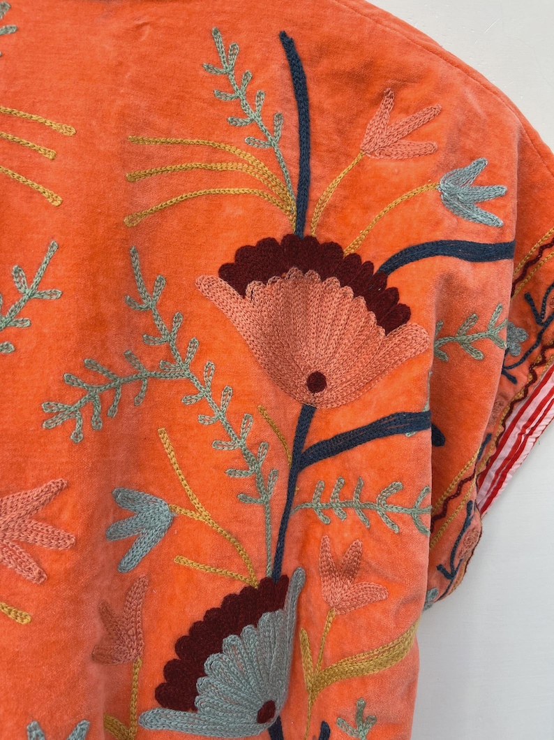 May include: Close-up of an orange embroidered garment. The garment features a floral design with pink, blue, and yellow thread. The embroidery is detailed and covers a large portion of the fabric. The garment's edge has a red and white striped trim.