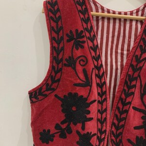 May include: A red velvet waistcoat with black embroidery of floral designs. The waistcoat has a scalloped hem and a V-neck.