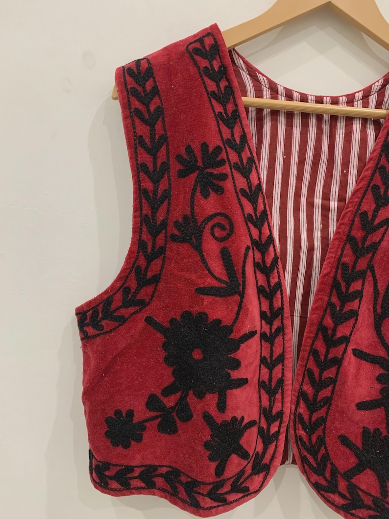 May include: A red velvet waistcoat with black embroidery of floral designs. The waistcoat has a scalloped hem and a V-neck.