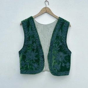 May include: A green velvet vest with floral embroidery and a striped lining. The vest is sleeveless and hangs on a wooden hanger against a white background. The embroidery is a darker shade of green.