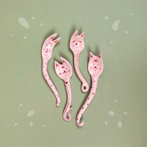 Könnte beinhalten: Vier rosa Keramik-Löffel mit katzenförmigen Köpfen und langen, gebogenen Griffen. Jeder Löffel hat ein bemaltes Katzengesicht mit gepunkteten Augen und herzförmigen Nasen. Die Griffe sind mit kleinen roten Herzen verziert.