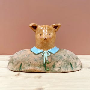 May include: A ceramic fox sculpture with a light brown head, blue collar, and a textured body. The fox has small, pointed ears and a sweet expression. The sculpture is a unique piece of art.