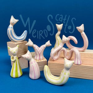 Puede incluir: Colección de figuras de gatos de cerámica caprichosas en varias poses. Los gatos presentan patrones de rayas en azul, rosa y amarillo. El texto "Weird Cats" está escrito en una fuente lúdica sobre las figuras. Los gatos se exhiben sobre pequeños bloques de madera sobre un fondo azul.
