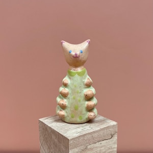 Könnte beinhalten: Eine skurrile Keramik-Katzenfigur. Die Katze hat einen hellbraunen Kopf mit rosa Ohren und blauen Augen. Sie trägt ein hellgrünes Kleid mit braunen Akzenten und einem grünen Kragen. Die Figur wird auf einem kleinen Holzblock vor einem rosa Hintergrund präsentiert.