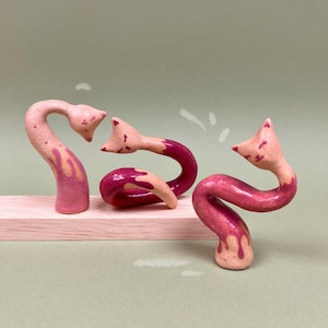 Puede incluir: Tres esculturas de cerámica caprichosas en tonos rosa y burdeos, que se asemejan a criaturas estilizadas. Están dispuestas sobre una tabla de madera clara sobre un fondo verde apagado. Las esculturas tienen un efecto de pintura texturizada y goteada.