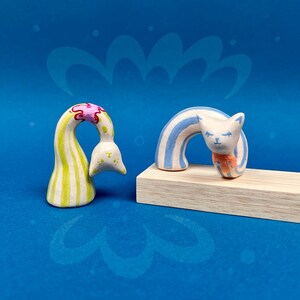 May include: Two whimsical ceramic sculptures on a blue background. One is a white and yellow striped form with pink and yellow accents. The other is a white cat with blue stripes and an orange scarf, posed on a wooden block.