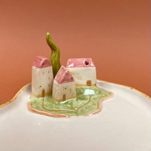 Può includere: Un piattino in ceramica con una scena di villaggio in miniatura scolpita. L'oggetto presenta tre piccole case con tetti rosa e pareti bianche, un alto albero verde e una base paesaggistica verde, il tutto su un vassoio curvo bianco sporco.