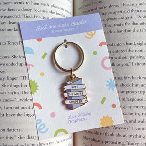 May include: A gold-coloured metal keyring with a charm featuring a stack of blue books and the words "Just One More Chapter." The charm has a white banner with the text. The keyring is on a light purple card with colourful confetti.