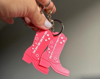 May include: Two bright pink cowboy boot keychains attached to a silver metal ring. Each boot features white raised lettering spelling out the names Hannah and Ashleigh, decorated with small white star shapes scattered across the surface.