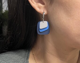 May include: A silver hoop earring with a layered, square pendant. The pendant has three layers: a dark blue base, a light blue middle layer, and a white top layer. The earring is a modern, geometric design.