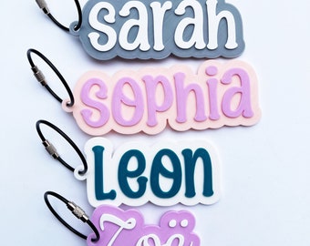 May include: Four personalised keyrings with the names "Sarah", "Sophia", "Leon", and "Zoë" written in white on colourful backgrounds. The keyrings are attached to black split rings.