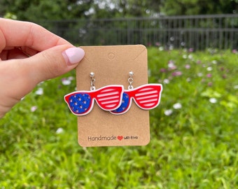 May include: A pair of red, white and blue American flag-themed sunglasses dangle earrings. The earrings are on a brown card with the text "Handmade with love".