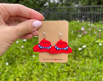 May include: A pair of red and blue earrings shaped like a hat with white stars. The earrings are on a brown card with the text "Handmade with love".