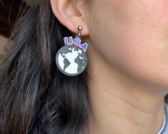 May include: A dangle earring featuring a circular charm with a black and white map of the world. Above the globe, the letters USA are printed in blue with a red outline. The earring is attached to a small silver-toned metal ball stud post.