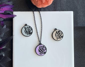 May include: Three round pendant necklaces with black chains, each featuring a letter and bat silhouettes. The letters are V, S, and A, with the S pendant having a lavender background. The pendants are on a white square surface, with orange velvet pumpkins in the background.