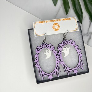 May include: A pair of Halloween-themed earrings featuring a lavender oval frame with a white ghost charm. The frame has a black outline and decorative elements. The earrings are displayed in a black box with the text "PRETTY PRINTED THINGS".