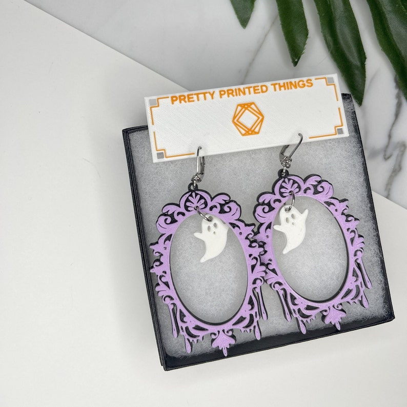 May include: A pair of Halloween-themed earrings featuring a lavender oval frame with a white ghost charm. The frame has a black outline and decorative elements. The earrings are displayed in a black box with the text "PRETTY PRINTED THINGS".