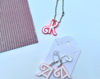 May include: A pink letter K necklace and a pair of pink letter A earrings. The earrings are on a white card with a silver hook.