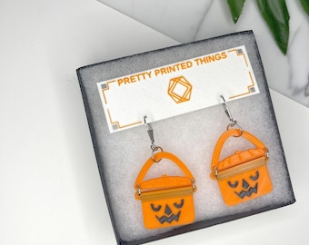 May include: Orange pumpkin-shaped earrings with black carved faces and silver-coloured ear wires. The earrings are presented in a black box with the text "PRETTY PRINTED THINGS" on a white label.