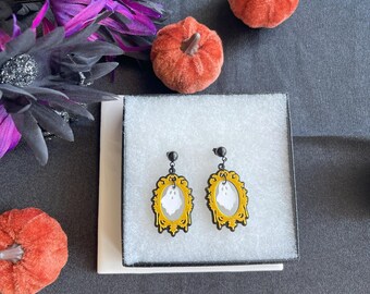 May include: A pair of Halloween earrings with a white ghost design in a yellow and black ornate frame. The earrings are presented in a white box, with orange pumpkins and purple and black floral decorations in the background.
