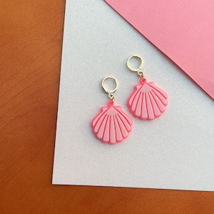 May include: A pair of pink seashell earrings with gold-colored hoops. The earrings are a light pink color with a darker pink outline and detailed shell design. The earrings are displayed on a white and pink background.