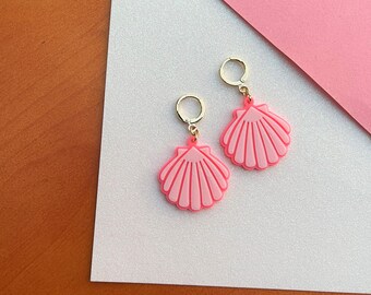 May include: A pair of pink seashell earrings with gold-colored hoops. The earrings are a light pink color with a darker pink outline and detailed shell design. The earrings are displayed on a white and pink background.