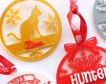 May include: Three personalized Christmas ornaments. One is gold with a cat silhouette and the name "Zion". Another is red with a snowflake design and the name "Hunter". The last is white with the text "2023 my Christmas".