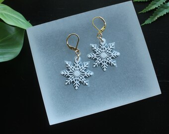 May include: A pair of white snowflake earrings with gold hooks. The snowflakes are delicate and intricate.