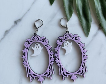 May include: A pair of Halloween earrings. Each earring has a light purple oval frame with a black filigree design and a small white ghost charm. The earrings have silver-coloured lever-back closures.