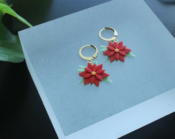 May include: A pair of gold hoop earrings with red poinsettia flower charms. The flowers have a yellow centre and green leaves.