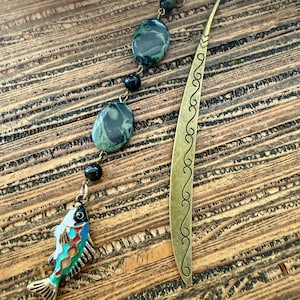 May include: A metal bookmark with an antique brass finish featuring a curved top and an engraved scroll pattern. A dangling chain attached to the top holds two dark green marbled oval beads, two small black round beads, and a colorful fish charm.