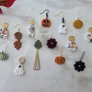 May include: A collection of fourteen individual dangle earrings featuring autumn and Halloween themes. Designs include pumpkins, ghosts, skulls, spider webs, flowers, and leaves crafted from clay and metal, suspended from gold or black earring hooks.