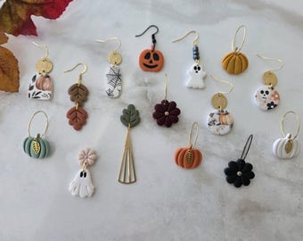Puede incluir: Una colección de catorce pendientes colgantes individuales con motivos de otoño y Halloween. Los diseños incluyen calabazas, fantasmas, calaveras, telarañas, flores y hojas hechas de arcilla y metal, suspendidos de ganchos dorados o negros.