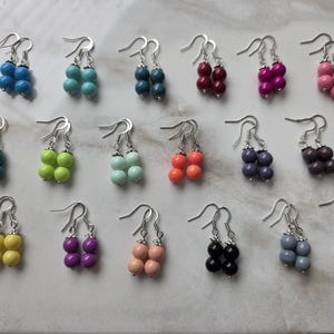 May include: A collection of seventeen pairs of drop earrings, each featuring two stacked round beads in various solid colors, including blue, teal, pink, red, purple, green, orange, yellow, black, gray, and white, attached to silver-toned metal hooks.