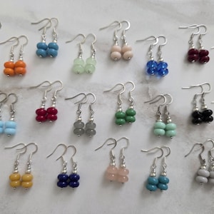 May include: A collection of colorful beaded dangle earrings. Each pair features two stacked, rounded beads in various colors, including white, orange, blue, green, and red. The earrings have silver-tone ear wires and are displayed on a light-colored surface.