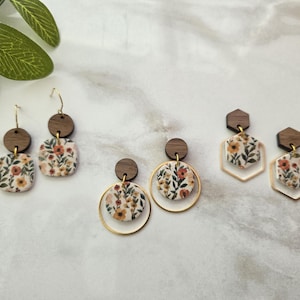 May include: A collection of floral earrings with wooden and gold accents. The earrings feature a variety of shapes, including squares, circles, and hexagons, with a cream-colored background and a floral pattern in shades of orange, yellow, and green.