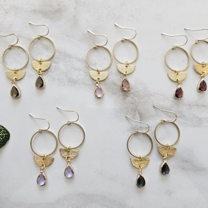 May include: Gold hoop earrings with a half-circle dangle and a teardrop-shaped gemstone. The earrings are available in three gemstone colors: purple, gray, and black.