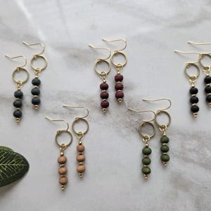 May include: A collection of handmade earrings with gold-colored hooks and circular accents. Each earring features three small, round beads in various colors, including black, burgundy, light brown, and olive green. The earrings are displayed on a white and gray marble surface.