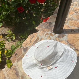 May include: White sun hat with a wide brim and cherry embroidery. The hat has a small label that says "Emily & Amelia".