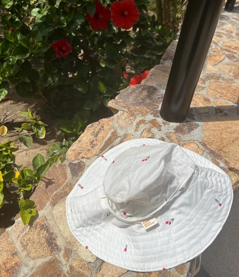 May include: White sun hat with a wide brim and cherry embroidery. The hat has a small label that says "Emily & Amelia".